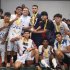Un grupo de estudiantes celebra su victoria en los Juegos Intercolegiados 2025 de Puente Aranda. Los jóvenes, vestidos con uniformes deportivos y medallas doradas en el cuello, posan sonrientes alrededor de un gran trofeo dorado que sostienen junto a dos adultos. La escena refleja alegría, unión y logro colectivo al finalizar la competencia deportiva.