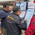 "Varias personas observan detenidamente una cartelera con información sobre proyectos y propuestas, durante una jornada de Presupuestos Participativos en la localidad de Puente Aranda. La actividad se realiza al aire libre, bajo una carpa, donde también hay un punto de atención con personal sentado y documentos visibles. Los ciudadanos participan activamente eligiendo iniciativas prioritarias para su comunidad."