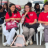 "Grupo de jóvenes sentados en sillas plásticas al aire libre durante un conversatorio juvenil en Puente Aranda. Al frente, tres jóvenes con camisetas rojas del programa 'MUSE' sonríen a la cámara, mientras otros participantes al fondo están atentos o revisan sus dispositivos. El ambiente es relajado, de diálogo y participación juvenil, en un espacio promovido por la Alcaldía Local y la Registraduría Nacional."